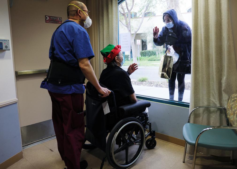 A person in a wheelchair waves to a person outside the window with face mask.