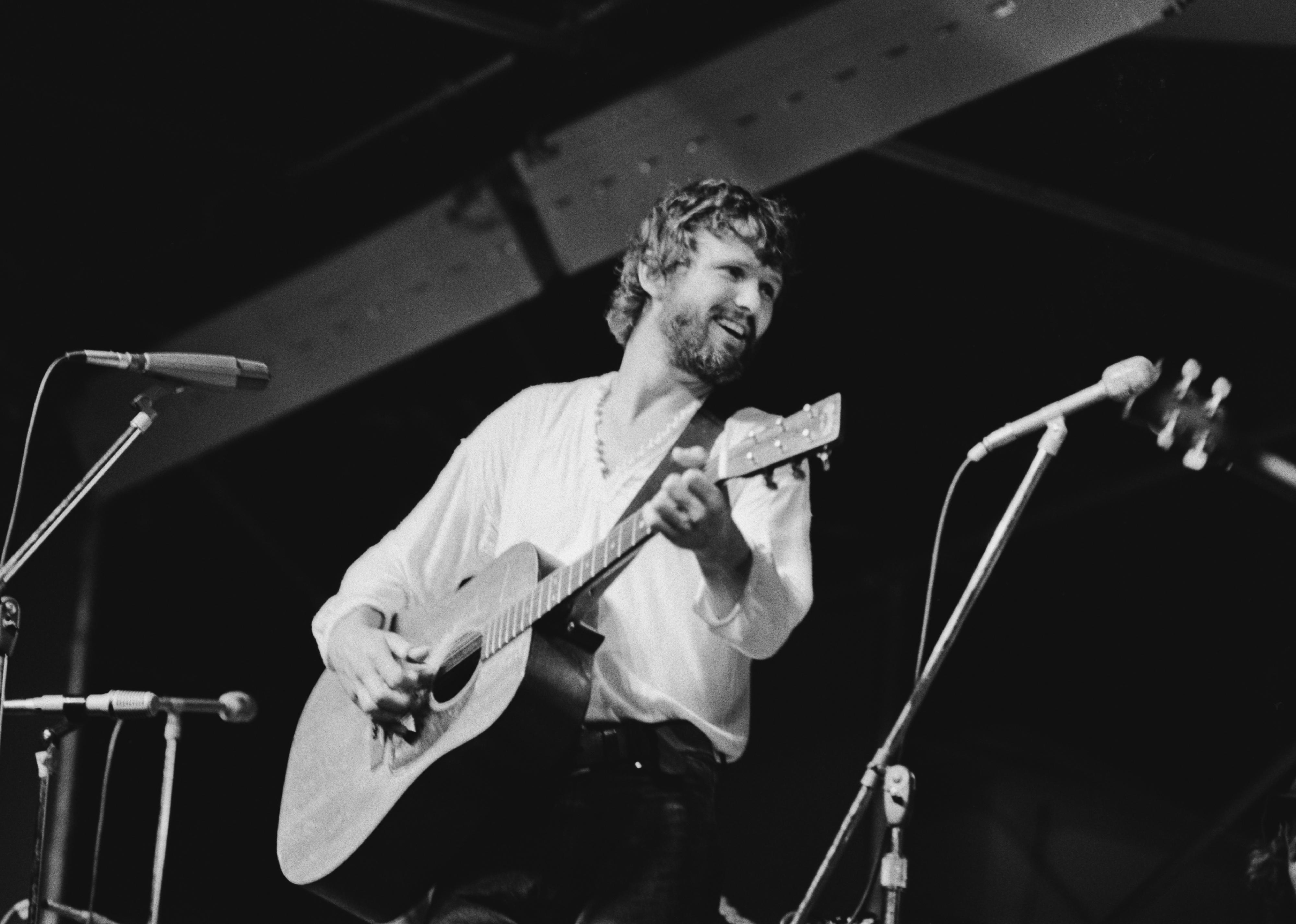 Kris Kristofferson, wearing a white v-neck top, playing an acoustic guitar.