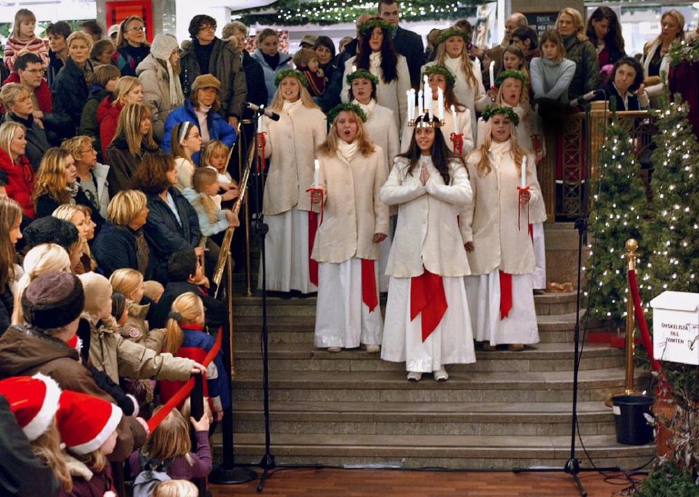 Crowds gather as several women dressed in white and red, holding candles, walk down a set of stairs. 