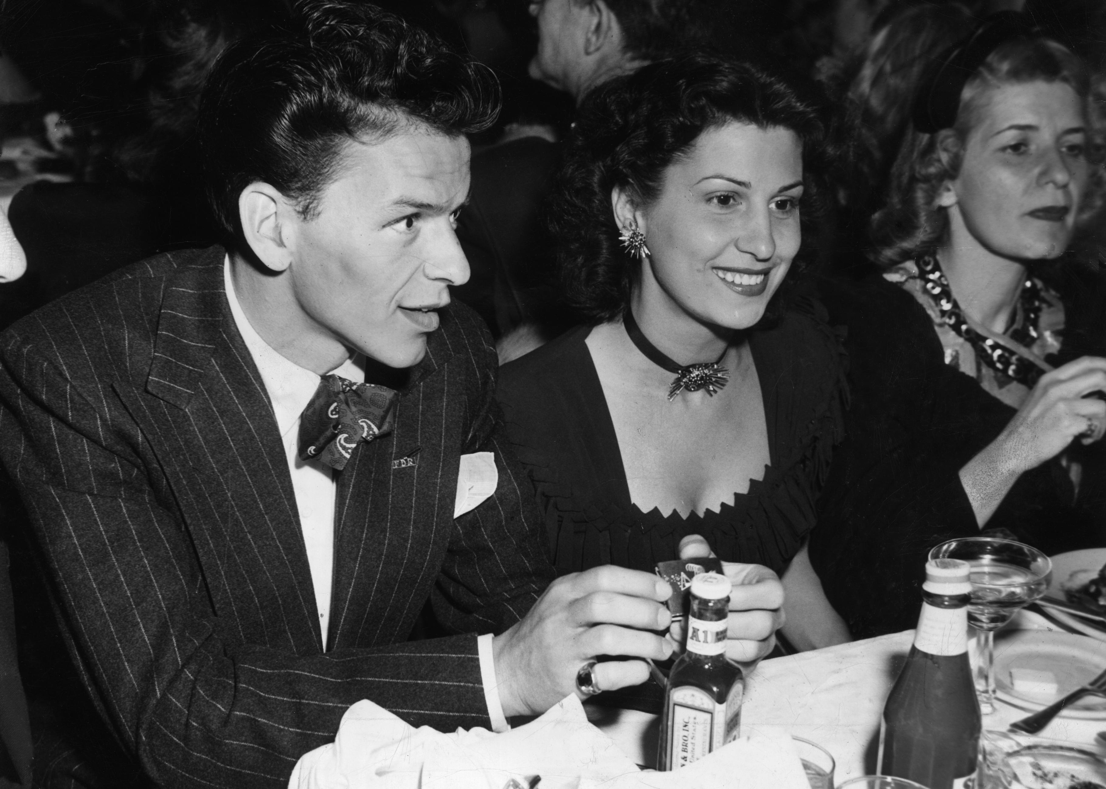 Frank Sinatra sitting at a table in a nightclub next to his first wife, Nancy Barbato. 