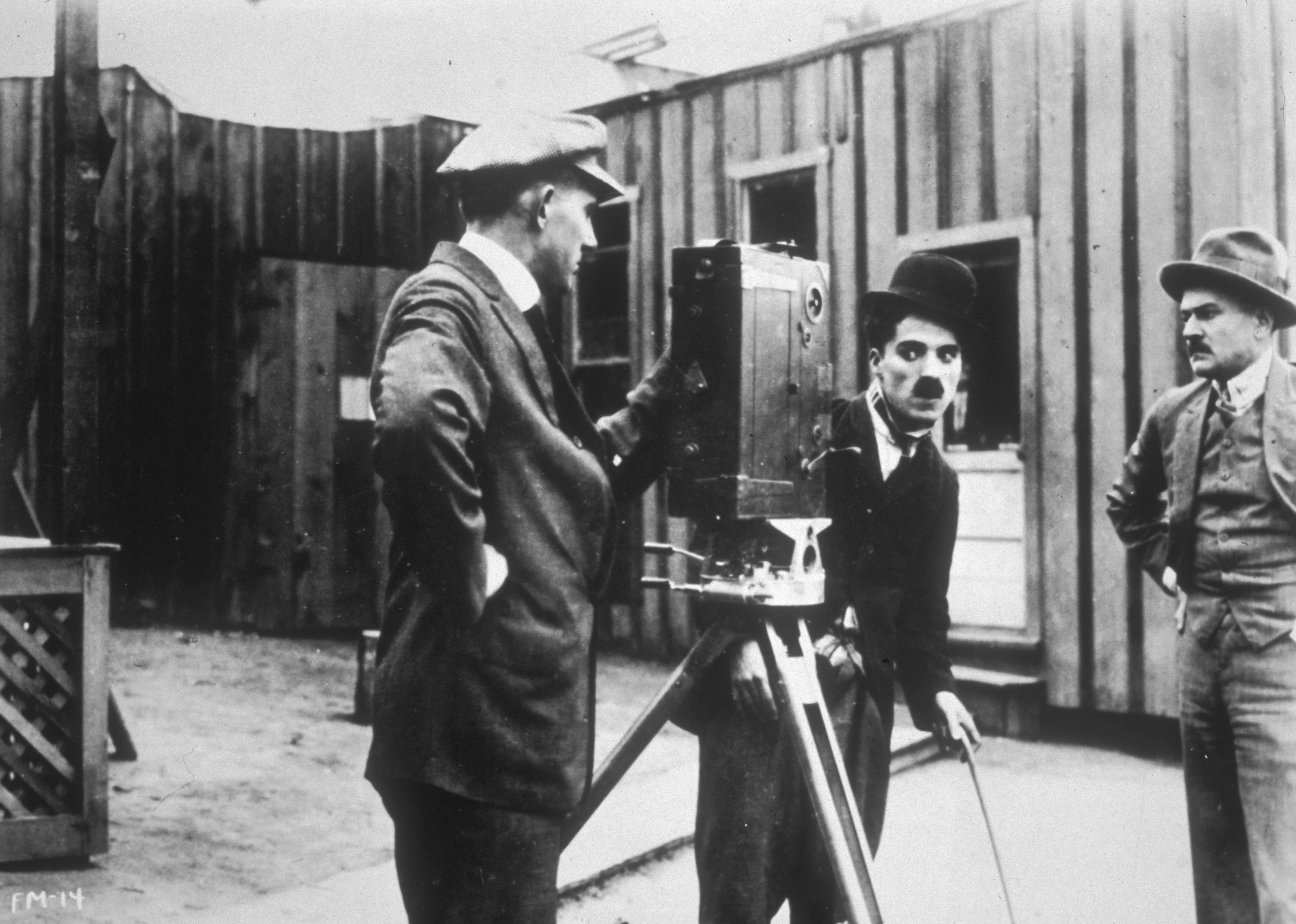 Charles Chaplin in costume as the Tramp, looks into a movie camera as two cameramen stand by.