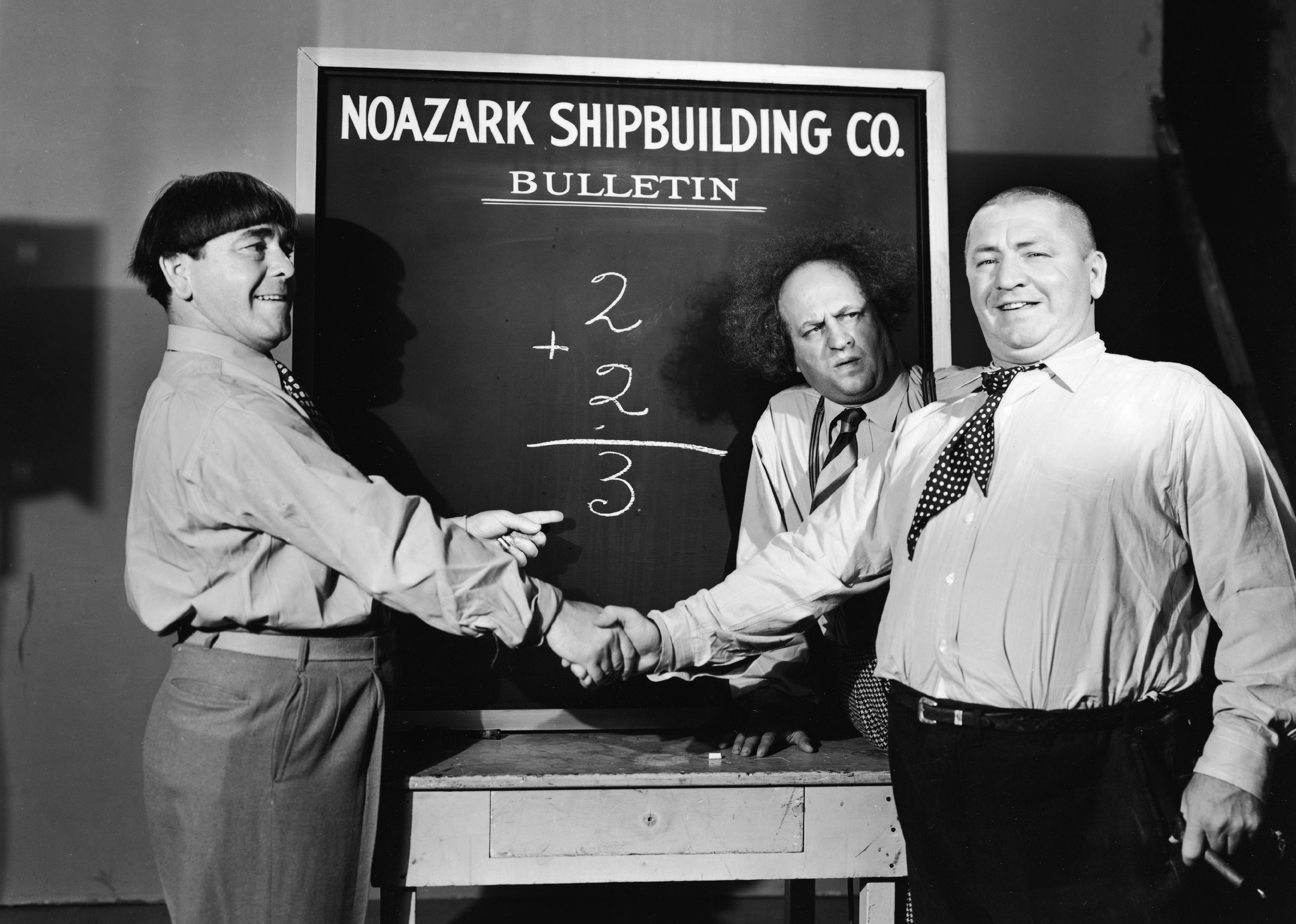 Moe Howard and Curly Howard shake hands as Larry Fine looks on in a still from a Three Stooges film. 