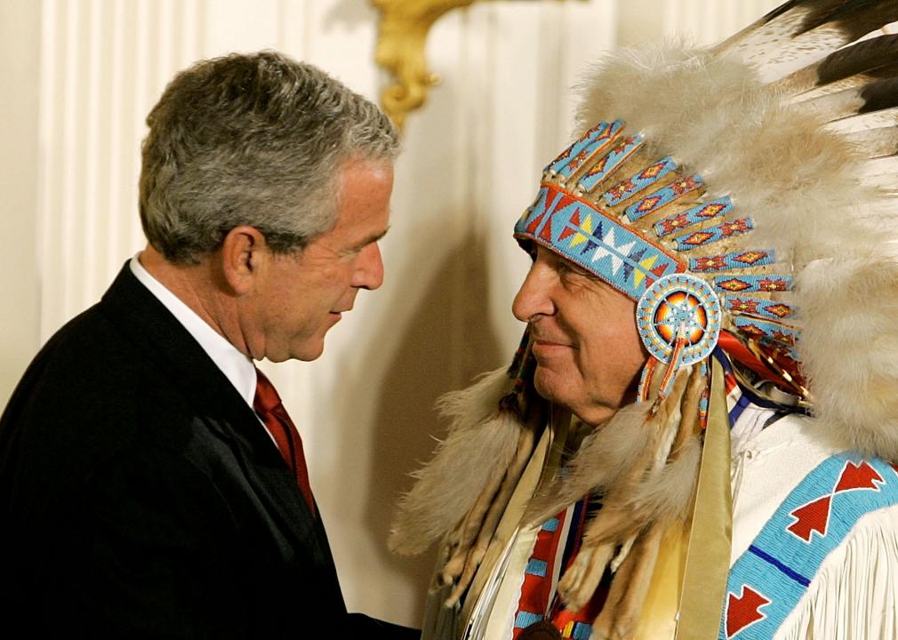Ben Nighthorse Campbell, wearing traditional Indigenous clothing, meets President George W. Bush. Both are gently smiling.