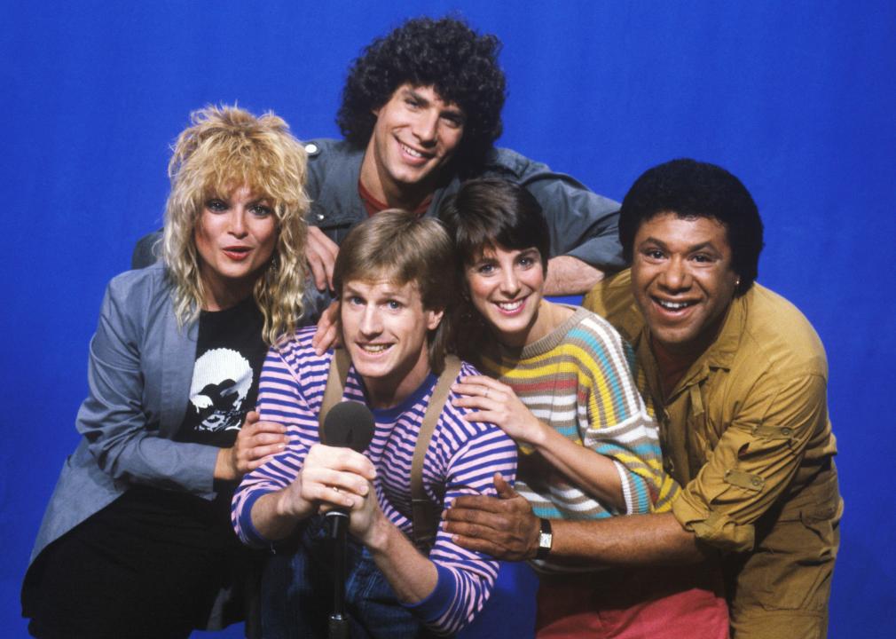 MTV VJs Mark Goodman, Nina Blackwood, Alan Hunter, Martha Quinn and JJ Jackson posing together against blue backdrop.