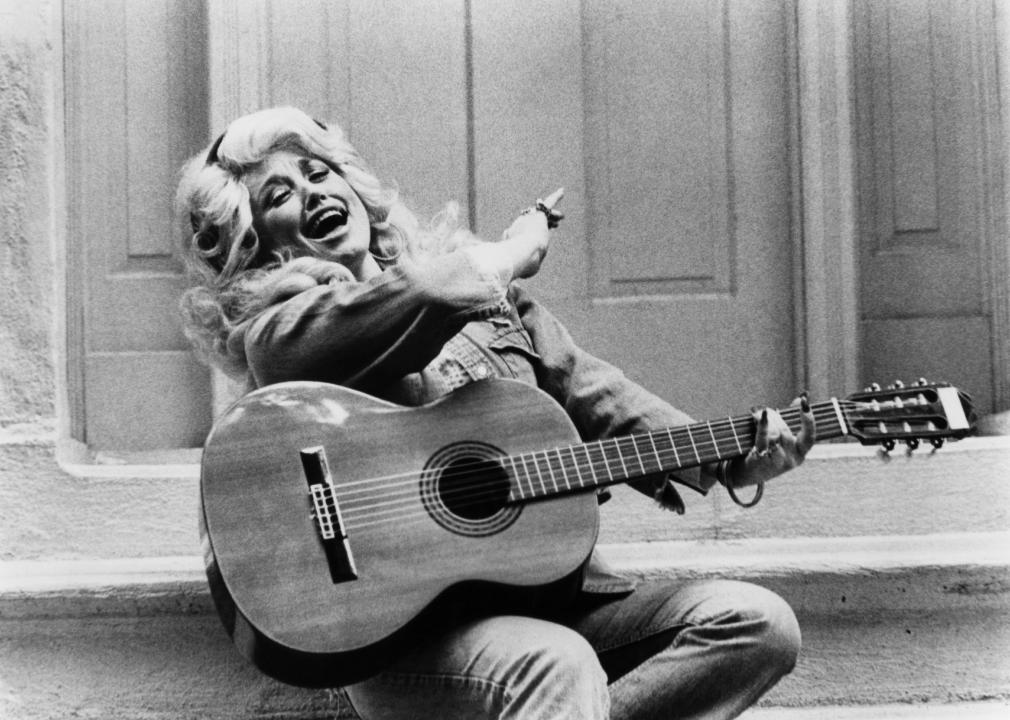 Dolly Parton posing on stoop with guitar in hand.