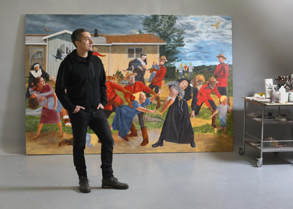 Kent Monkman stands in front of a very large painting of his called, The Scream, which shows Canadian police, priests, and nuns violently ripping Indigenous children from their parents to send them to residential schools.