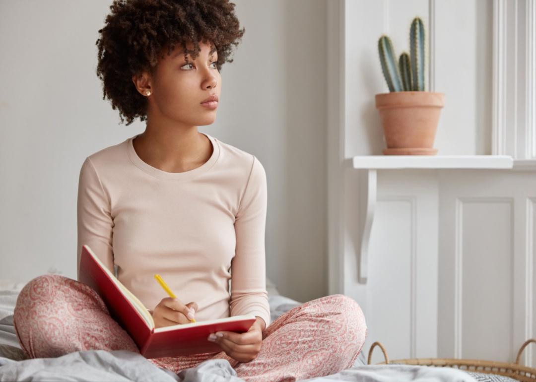 A person sits on a bed in pajamas writing and looking out the window.