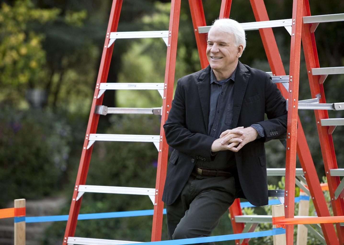 Steve Martin standing up against a ladder.