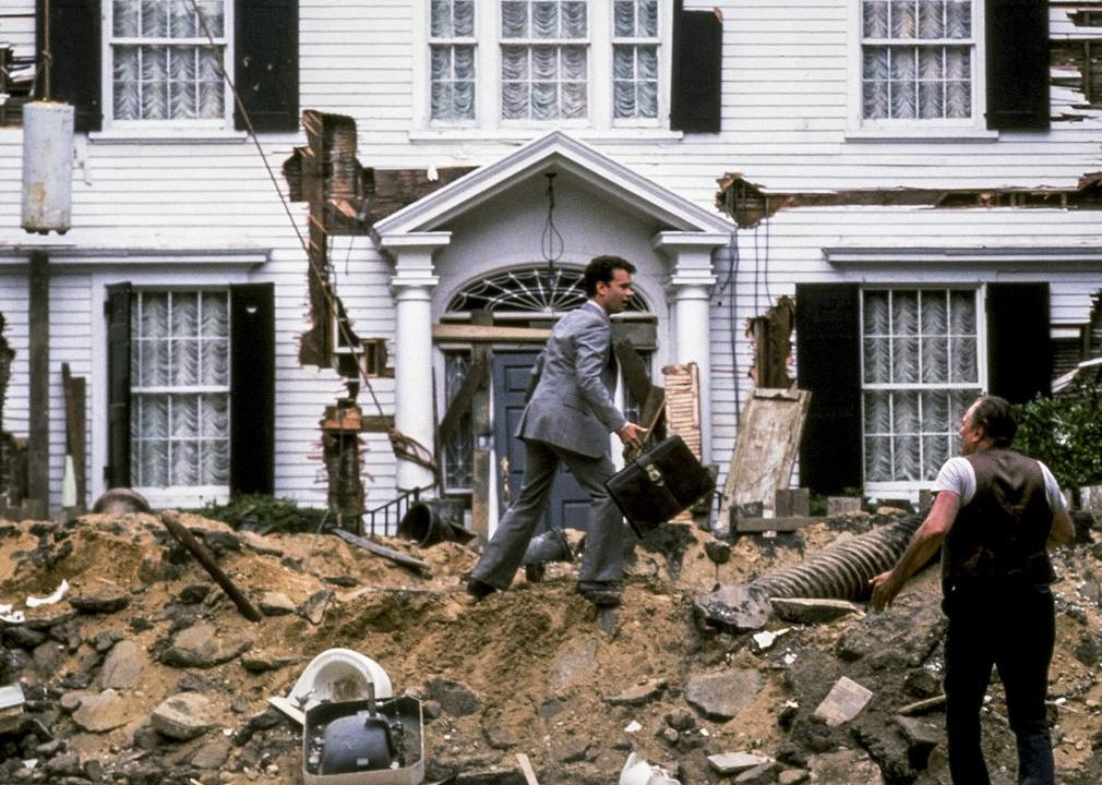 Tom Hanks in a suit in front of a torn-up lawn and house falling down.