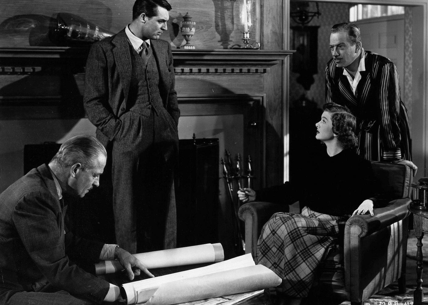 Cary Grant talks with a group of people in a living room with blueprints on a table.