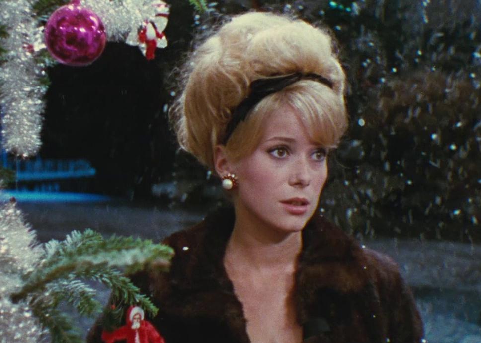Catherine Deneuve in a brown fur coat next to a Christmas tree.