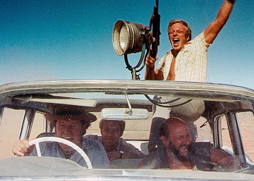 Four men laughing, yelling and waving guns in a moving car.