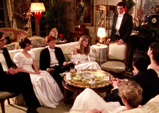 A group of guys in black suits and women in white dresses sitting on a couch in an elegant living room.