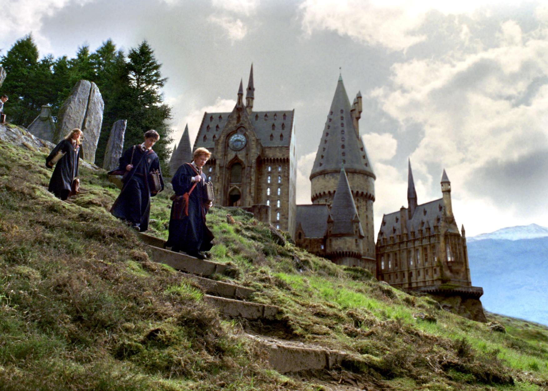 Rupert Grint, Daniel Radcliffe, and Emma Watson rush down the steps of a steep hill with a castle in the background.