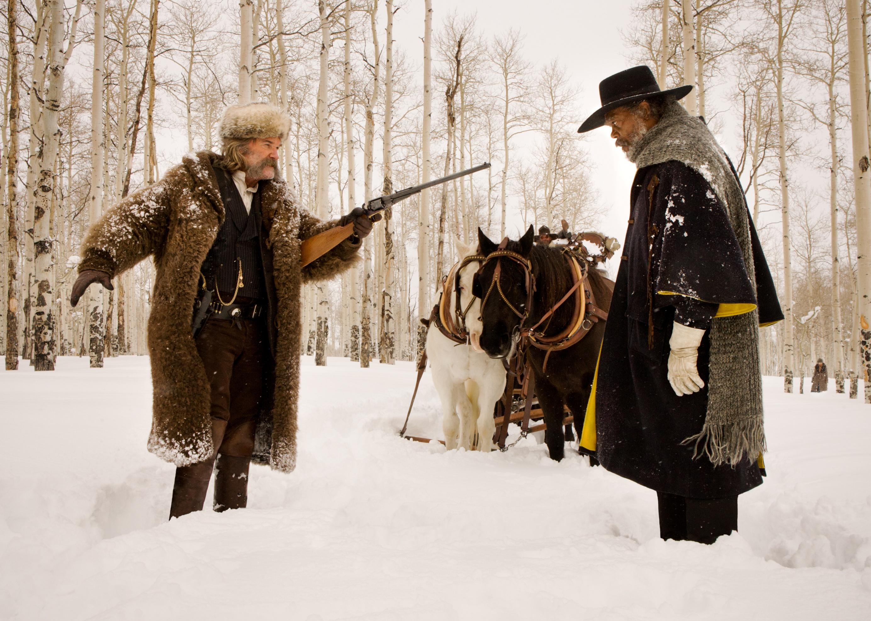 Kurt Russell points a long gun at Samuel L. Jackson with horses in the background in the snow.
