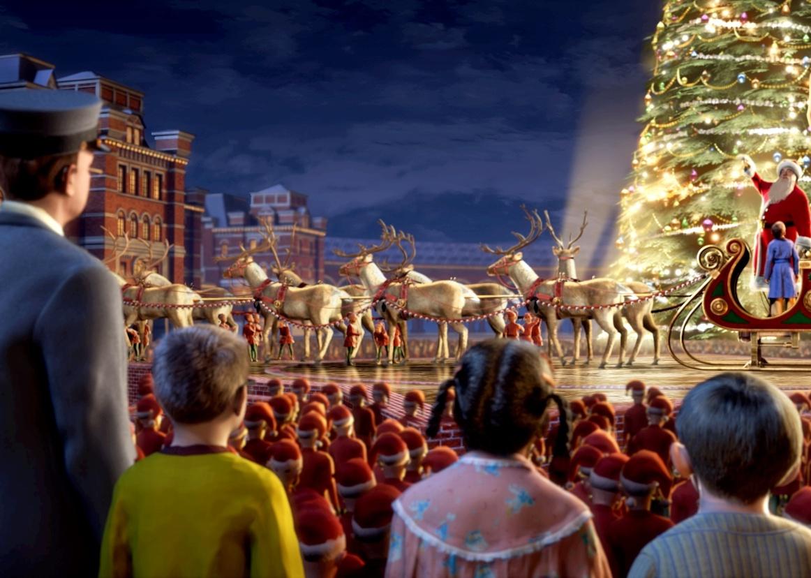 A crowd watches as a little boy sits in Santa's sleigh on a stage with a huge Christmas tree.