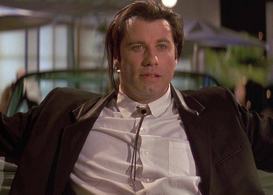 John Travolta with long hair sitting in the back of a convertible.