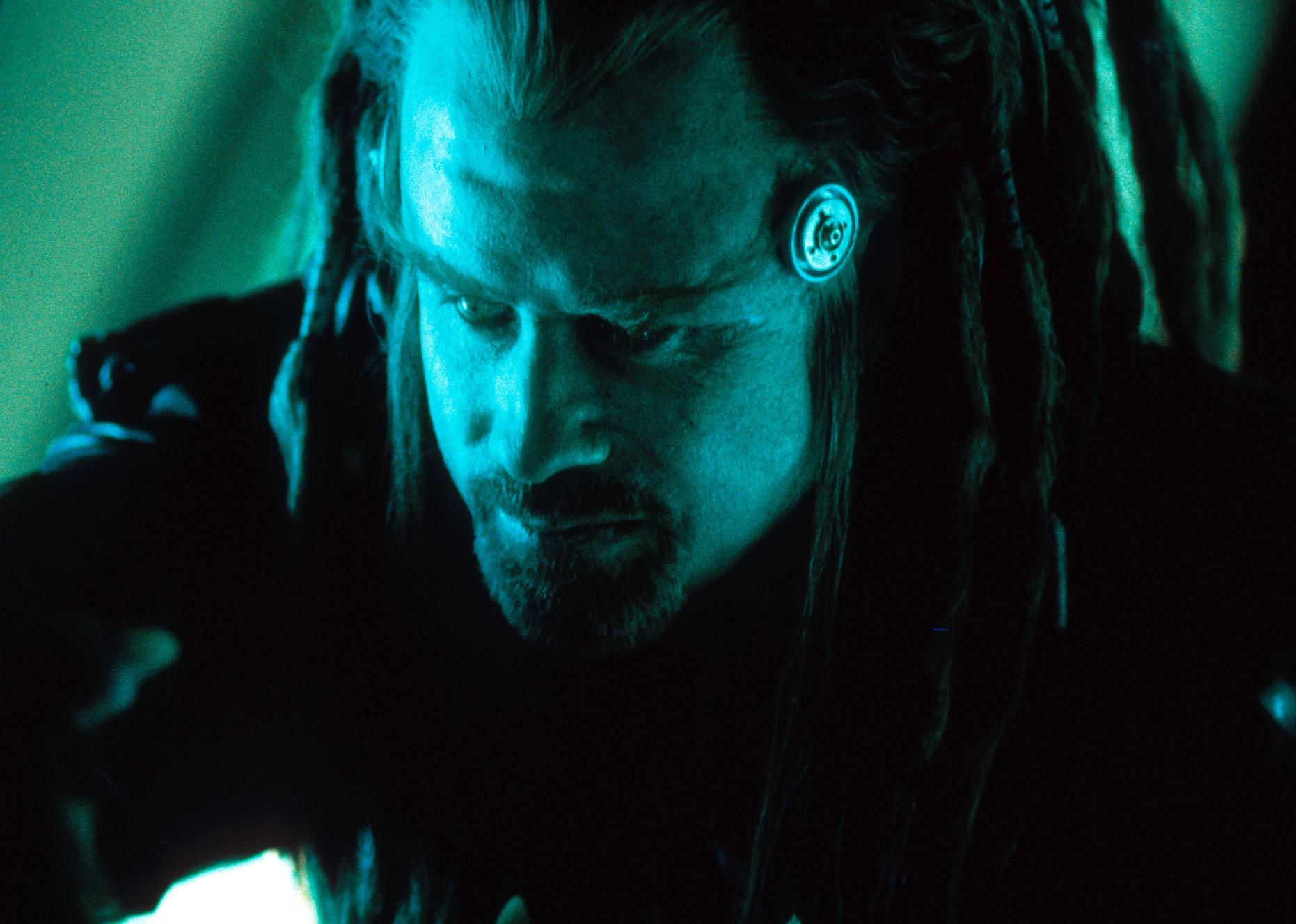 John Travolta under a green light with long dreadlocks and a round device on his temple.