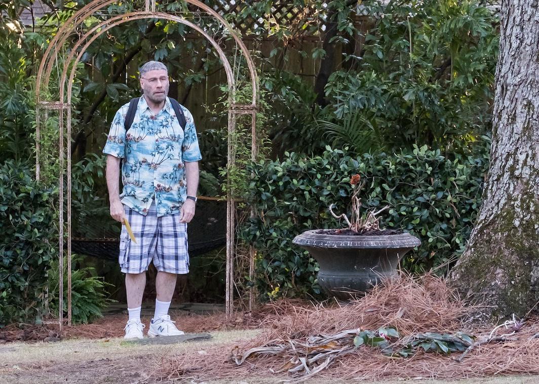John Travolta standing under an arch outside wearing plaid shorts and a hawaiian shirt.
