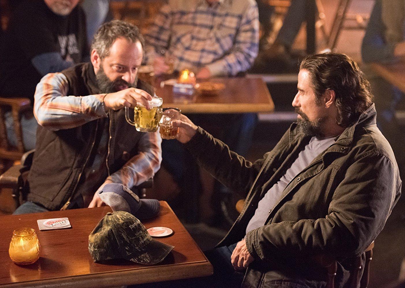 John Travolta and Gil Bellows at a tablein a bar cheersing with beers.
