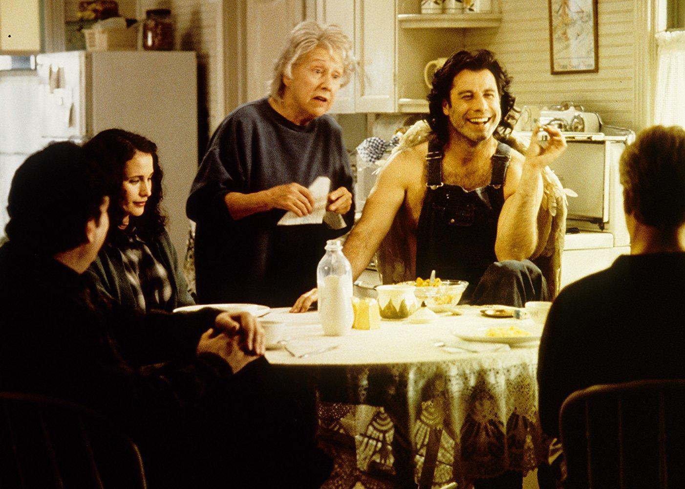 John Travolta, wearing overalls with no shirt, and Andie MacDowell sitting at a table with several other people.