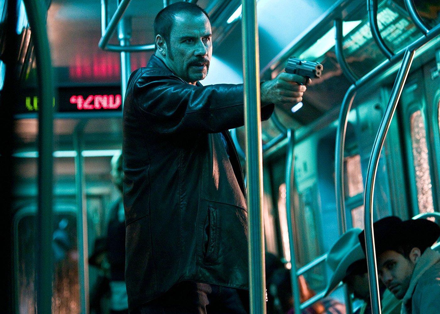 John Travolta, with short hair and a mustache, pointing a gun on a train.