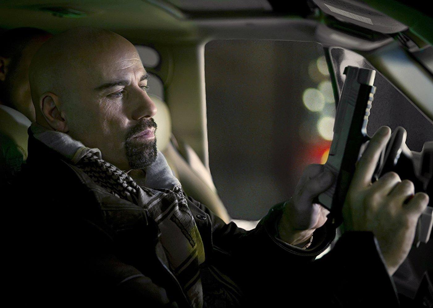 John Travolta, with a shaved head, holding a gun inside a vehicle.