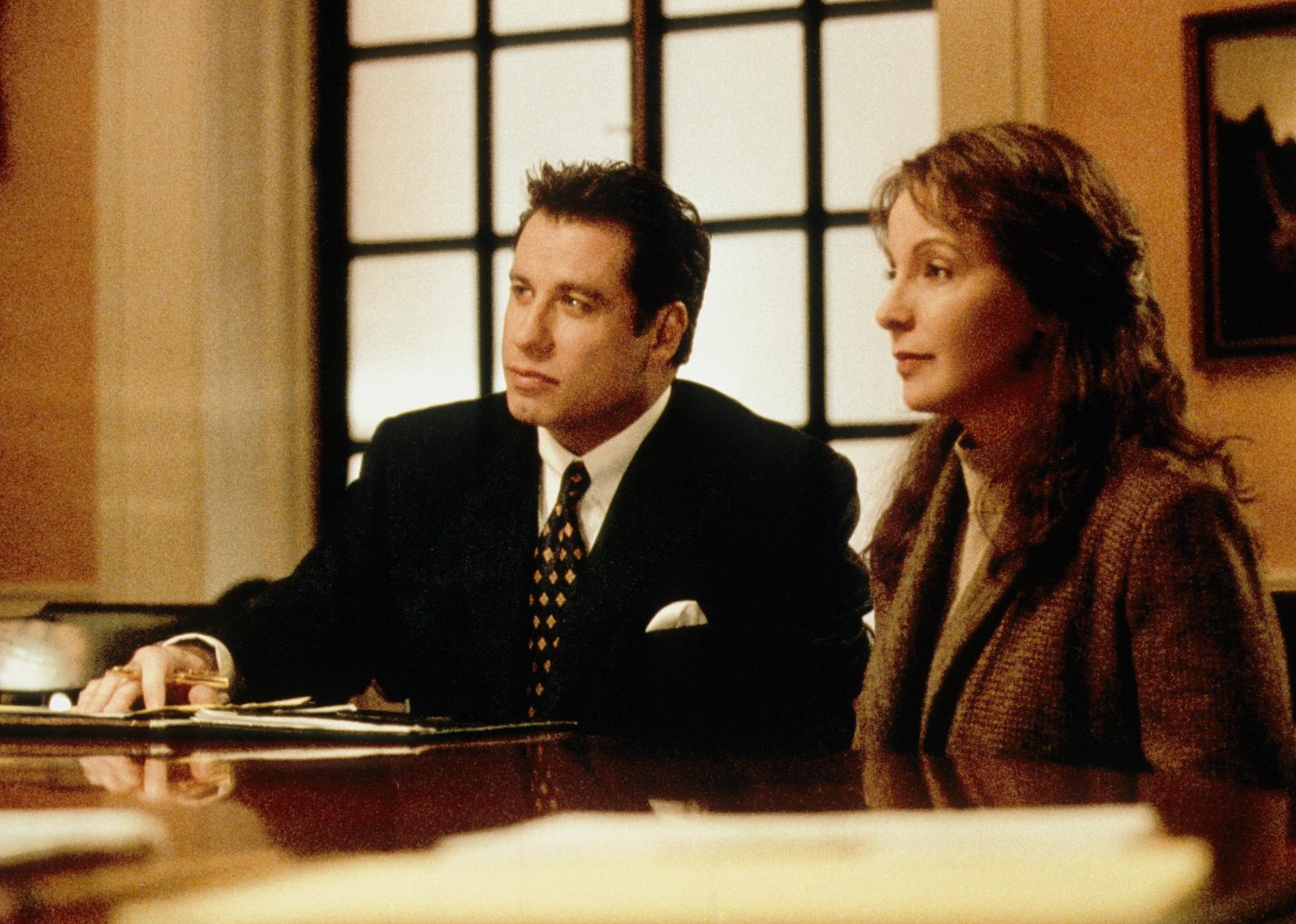 John Travolta in a suit talking to a woman in a coat inside a court room.