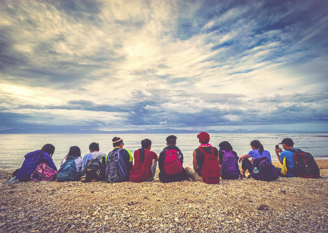Kids with backpacks sitting on a beach facing the ocean.
