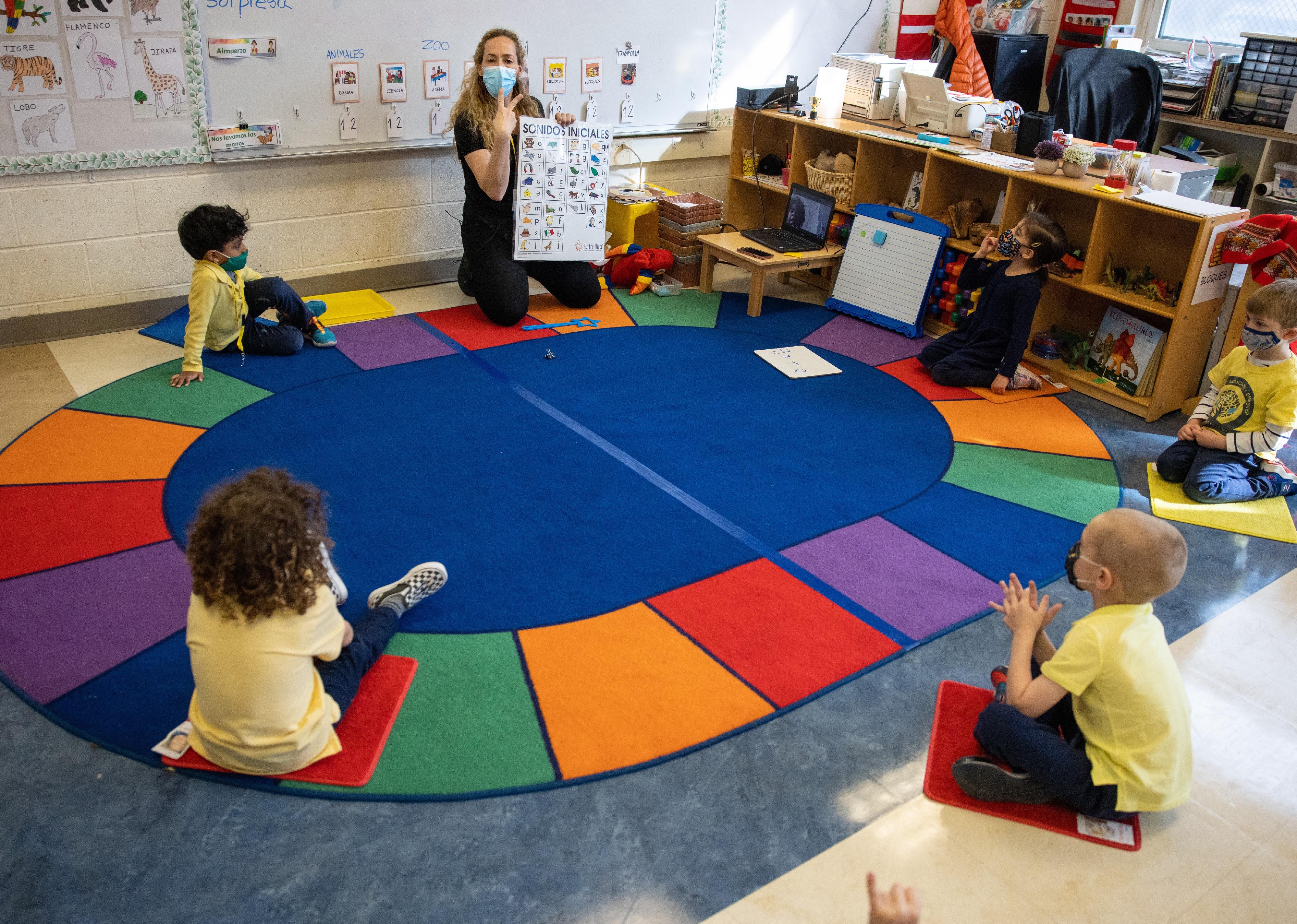 A teacher holding up a bilingual poster and using hand signals to communicate with children in a circle on the floor.