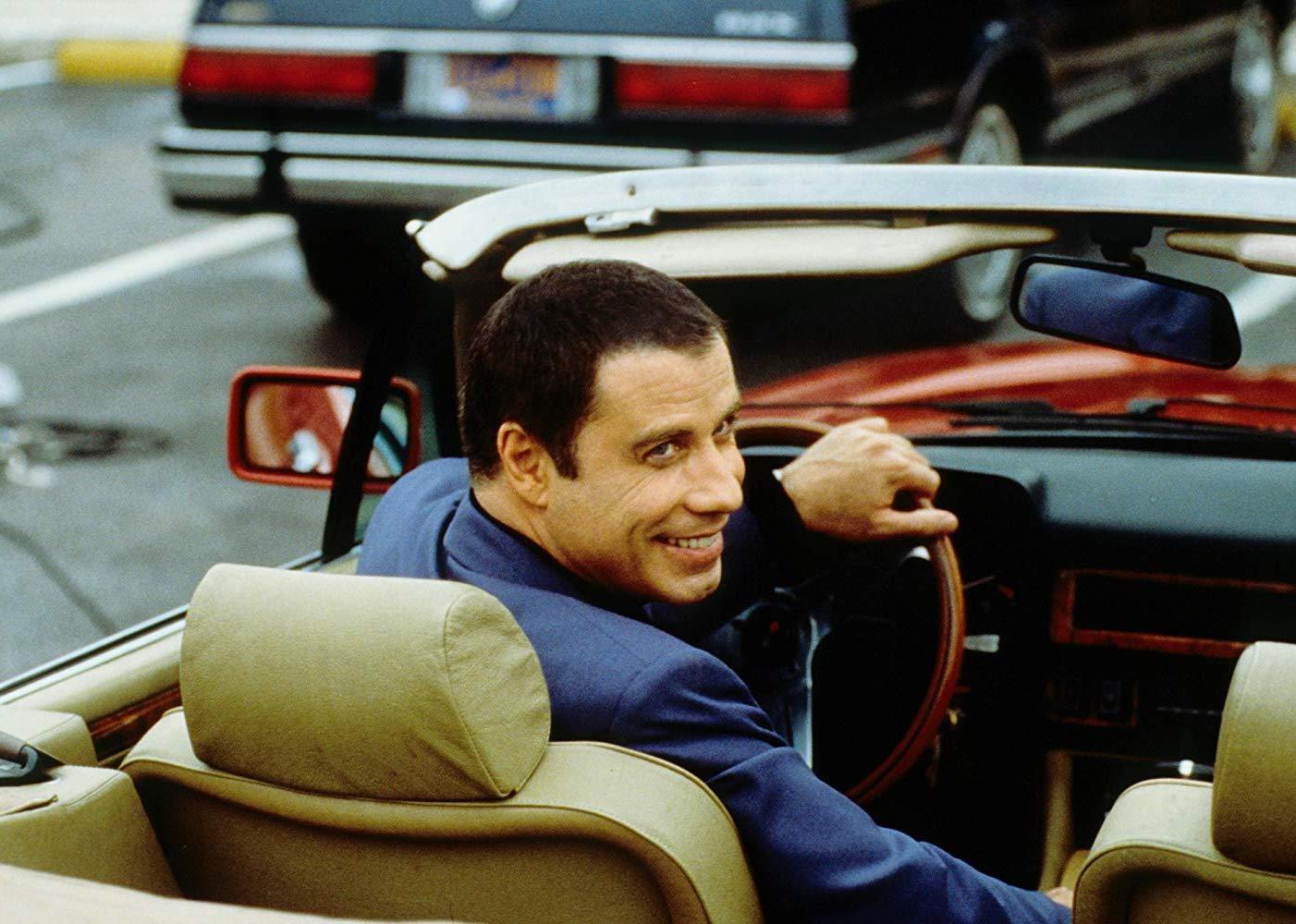 John Travolta smiles in the driver seat of a convertible