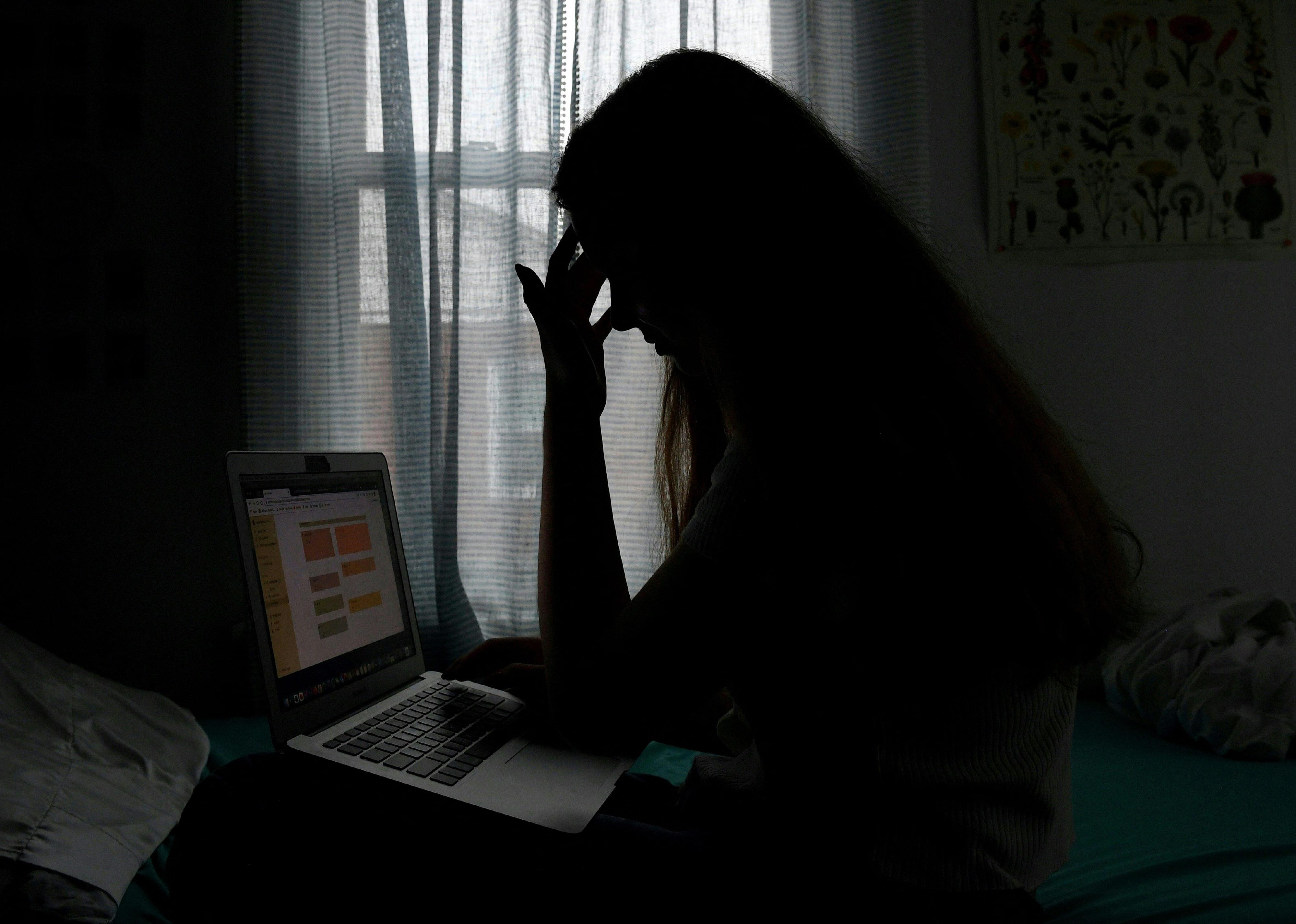 A teenager in the shadows sitting on a bed with her hand on her head.