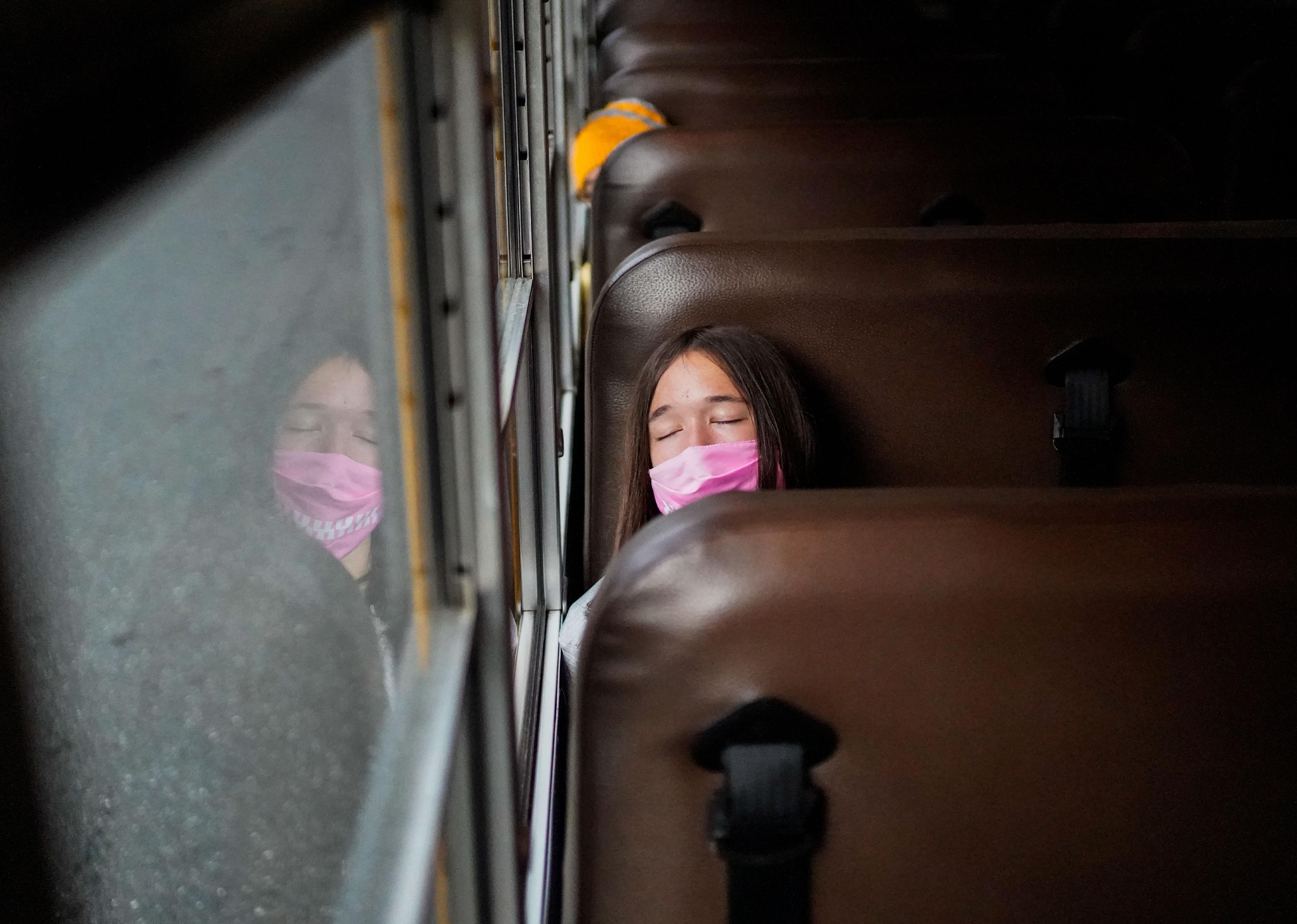 A teen riding the bus while resting their eyes and a wearing a pink mask.