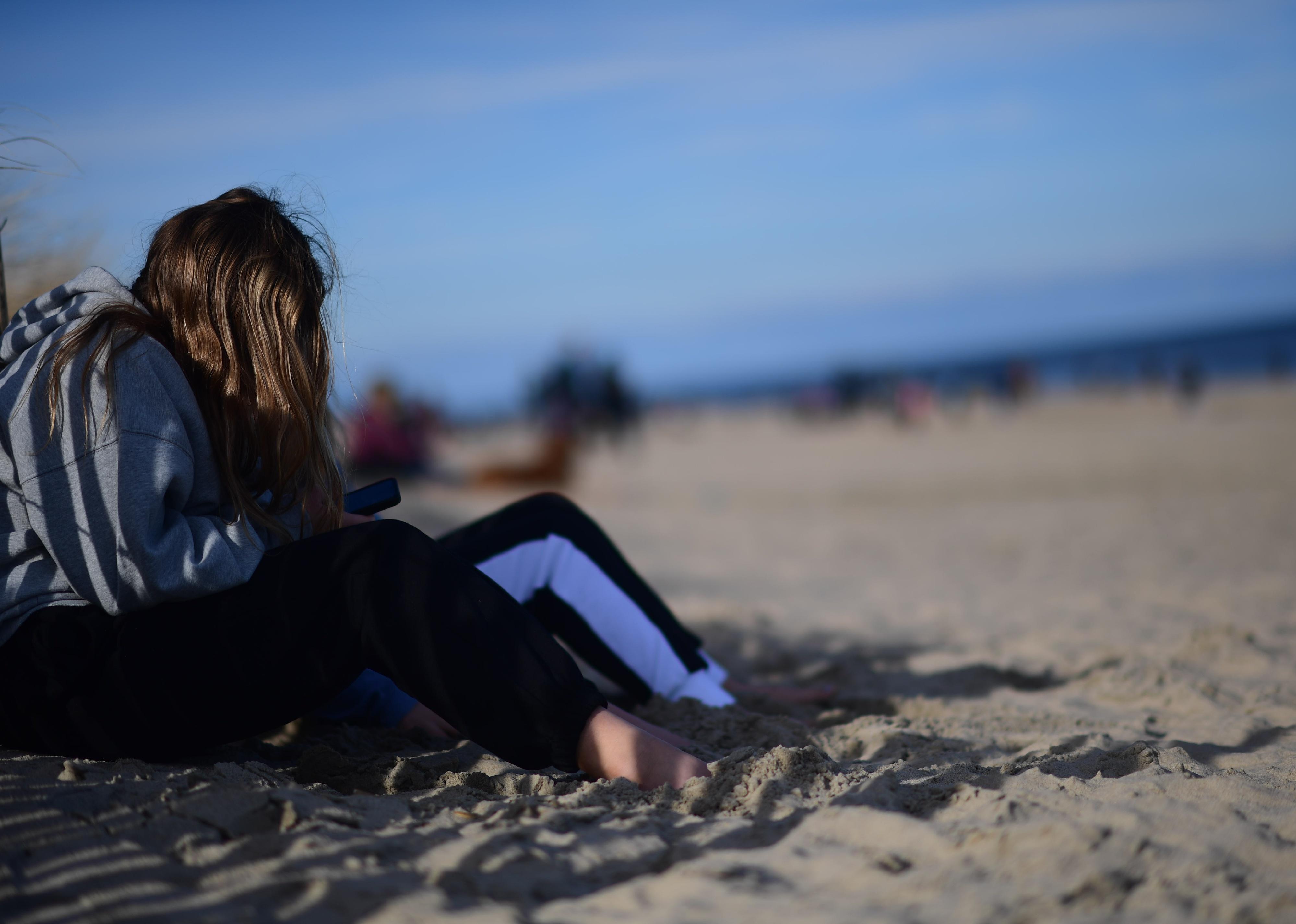 A teen sitting on the beach looking down at her phone.