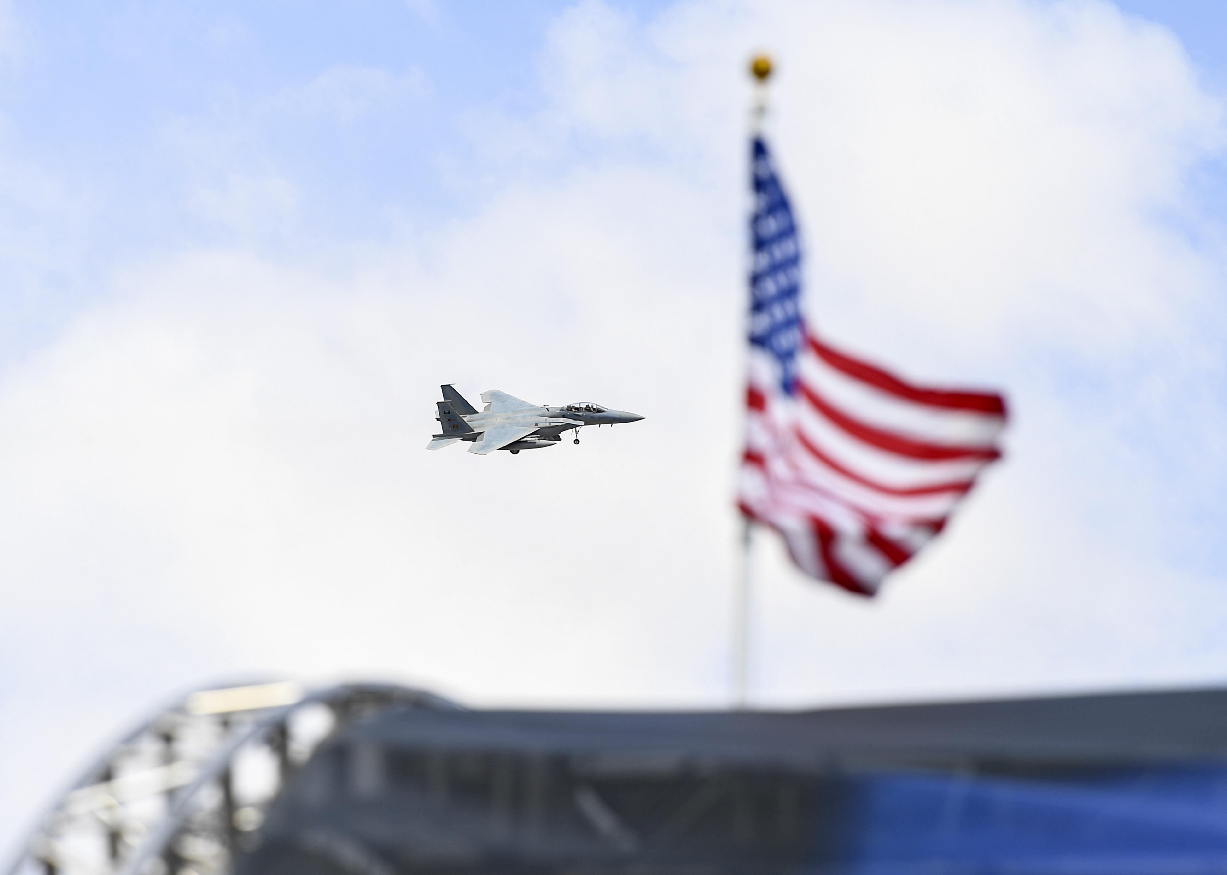 An F-15 approaching to land with an American flag in the foreground.