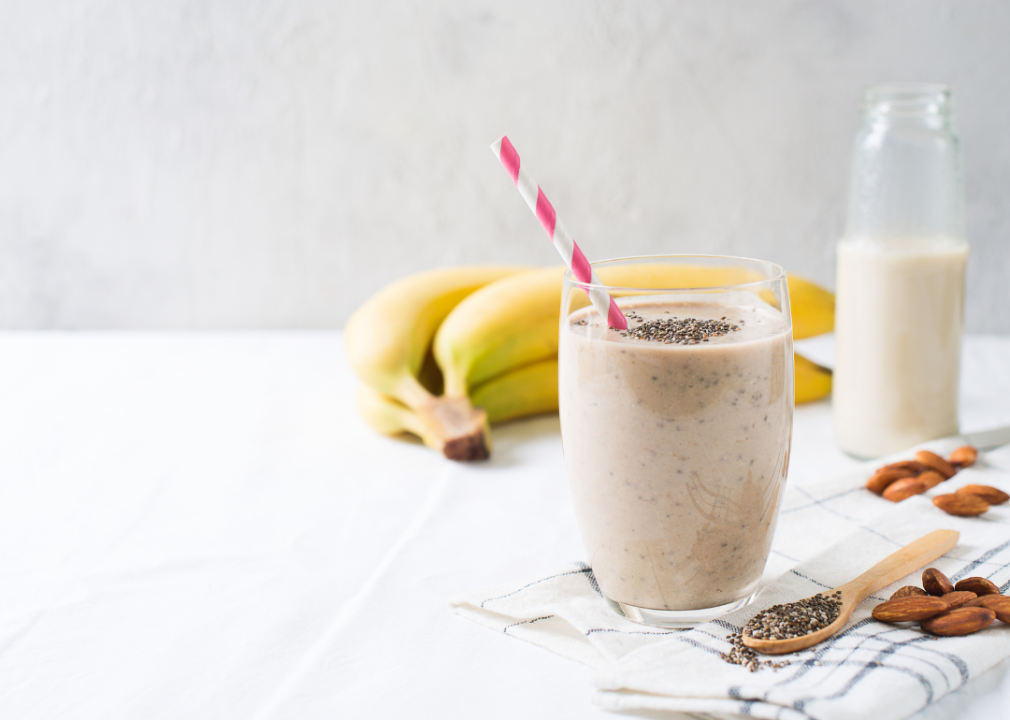 A smoothie on a table next to some bananas, seeds in a spoon, almonds and almond milk.