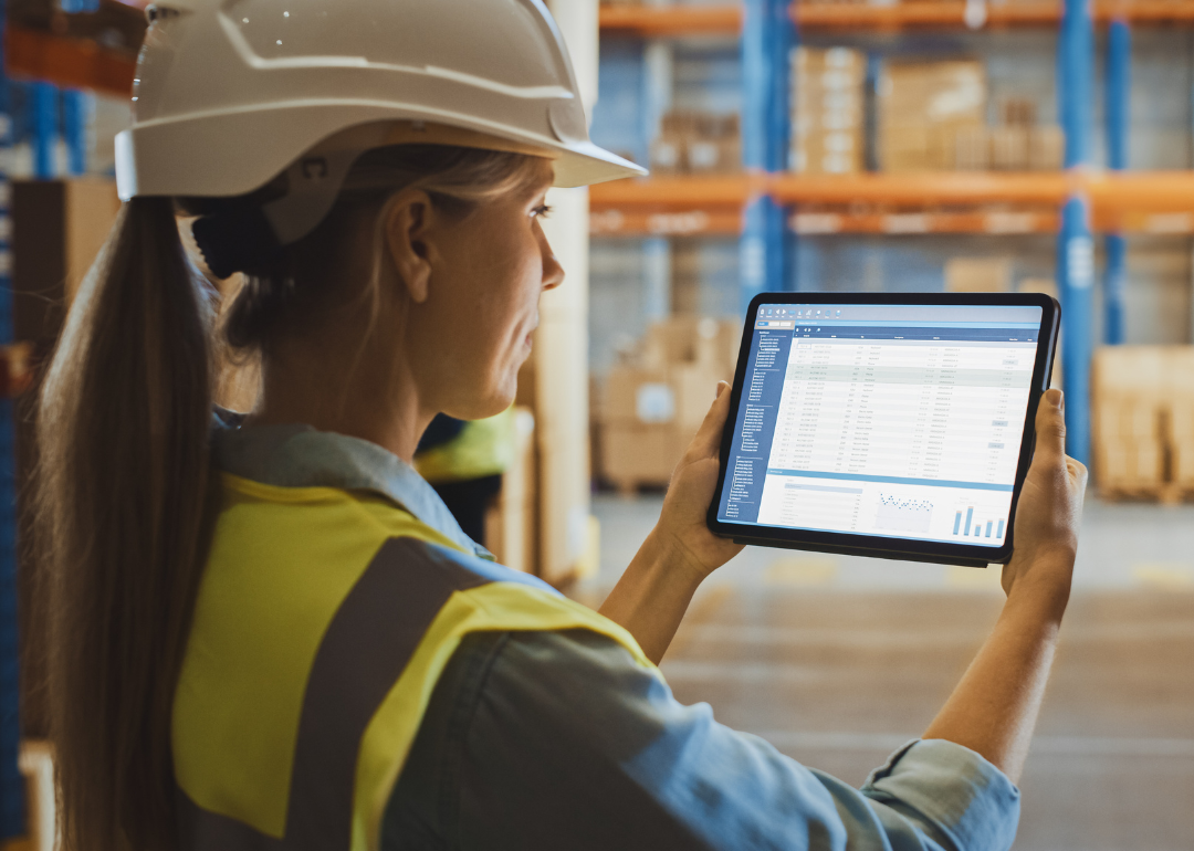 A woman wearing a hardhat in a warehouse looking at a spreadsheet on a tablet.