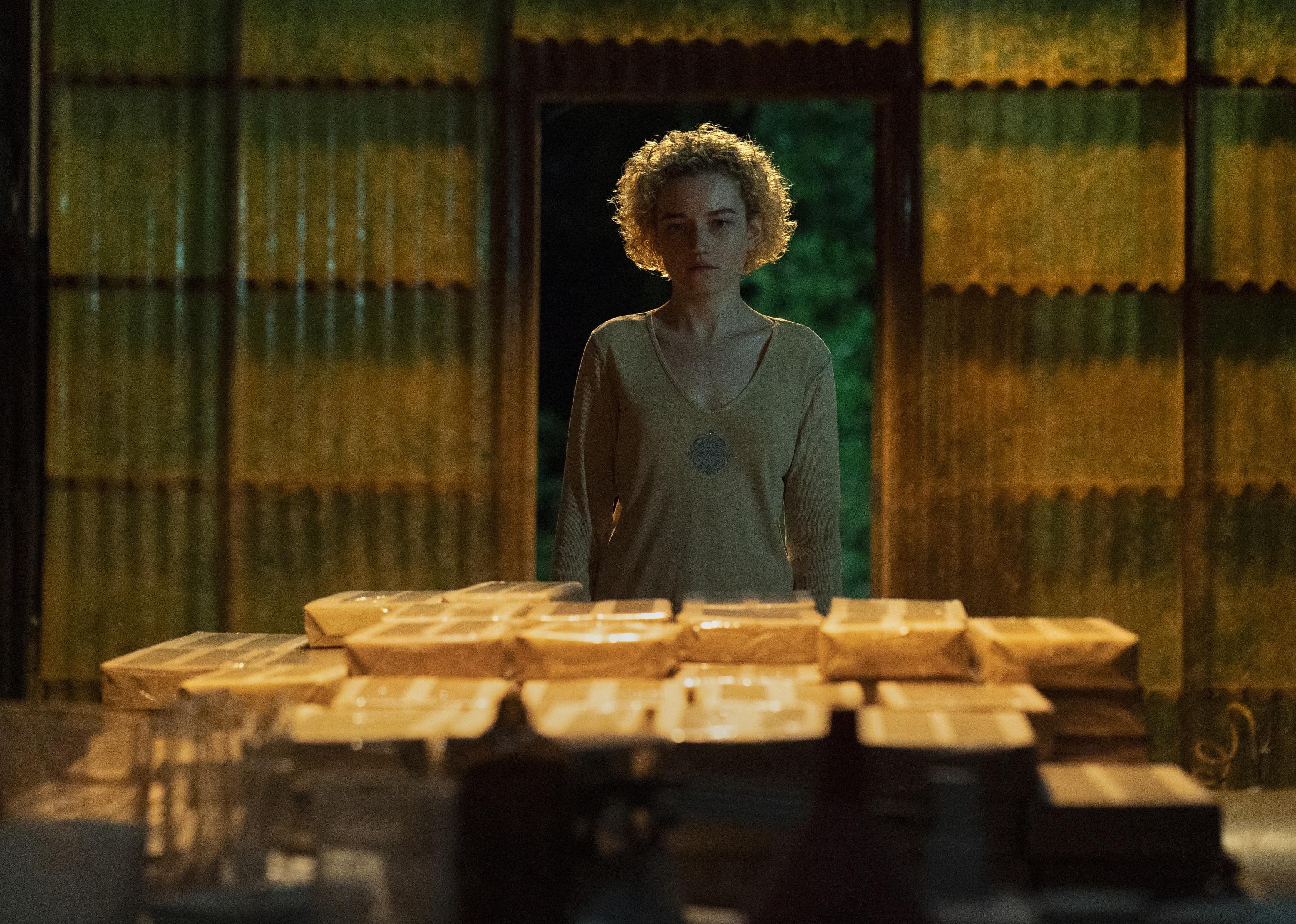 Julia Garner in a room looking at stacks of something wrapped in paper.