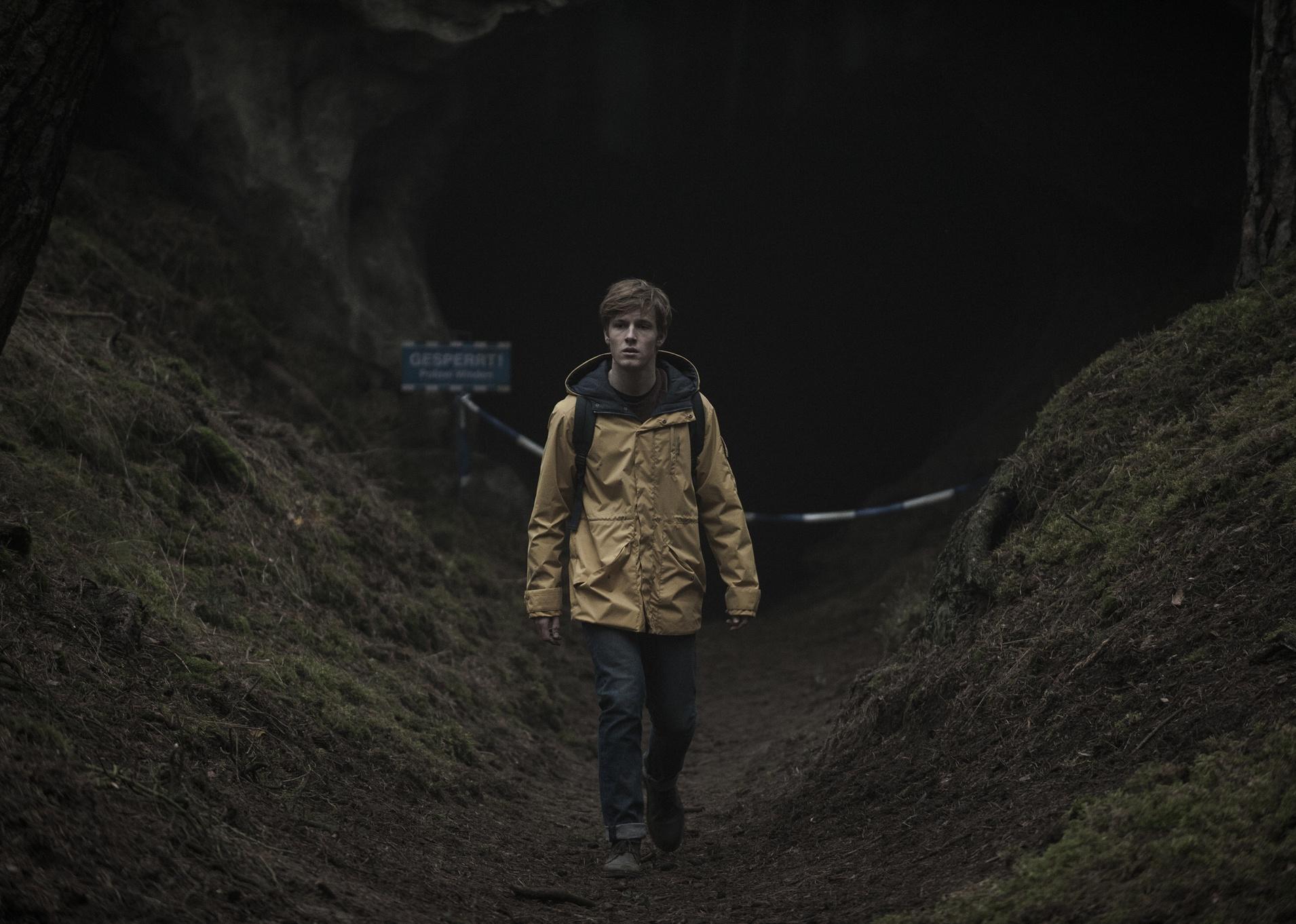 A boy in a yellow raincoat walking near a dark cave.