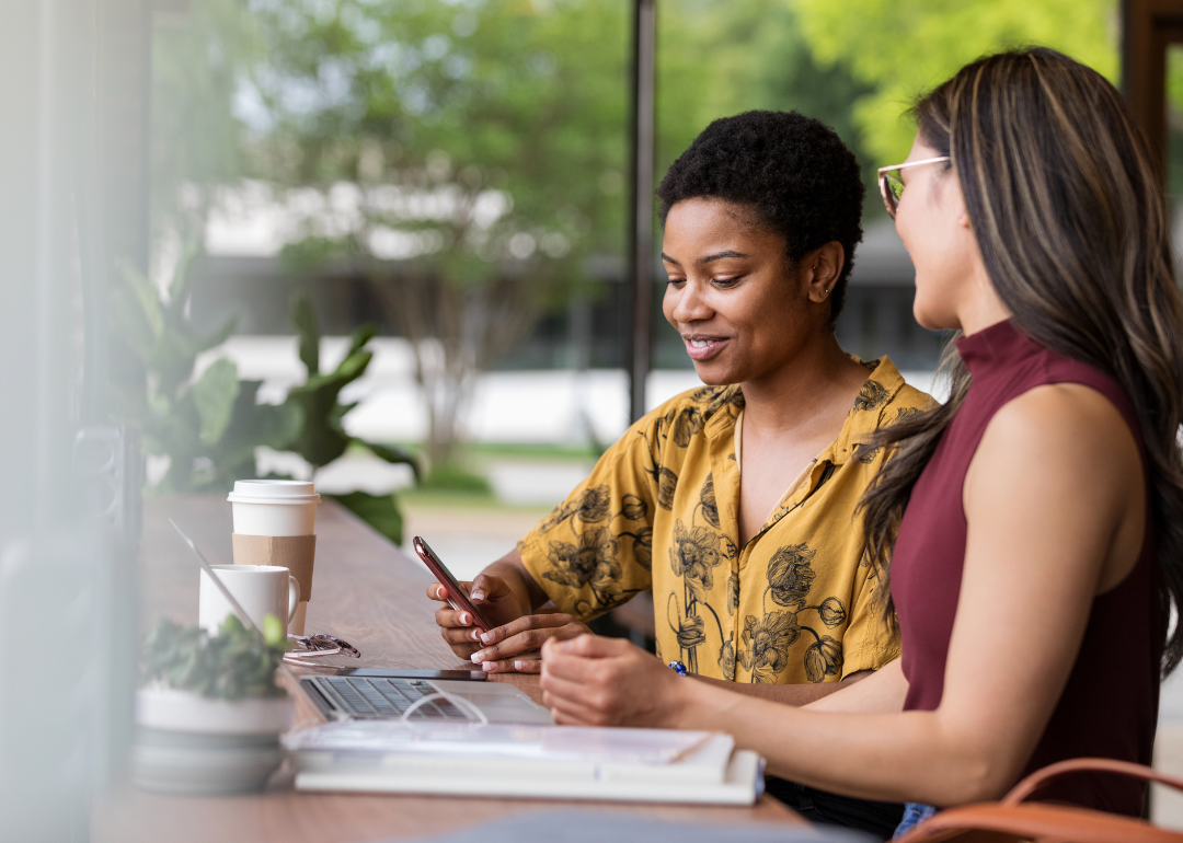 Two women at a laptop looking at a phone and smiling.