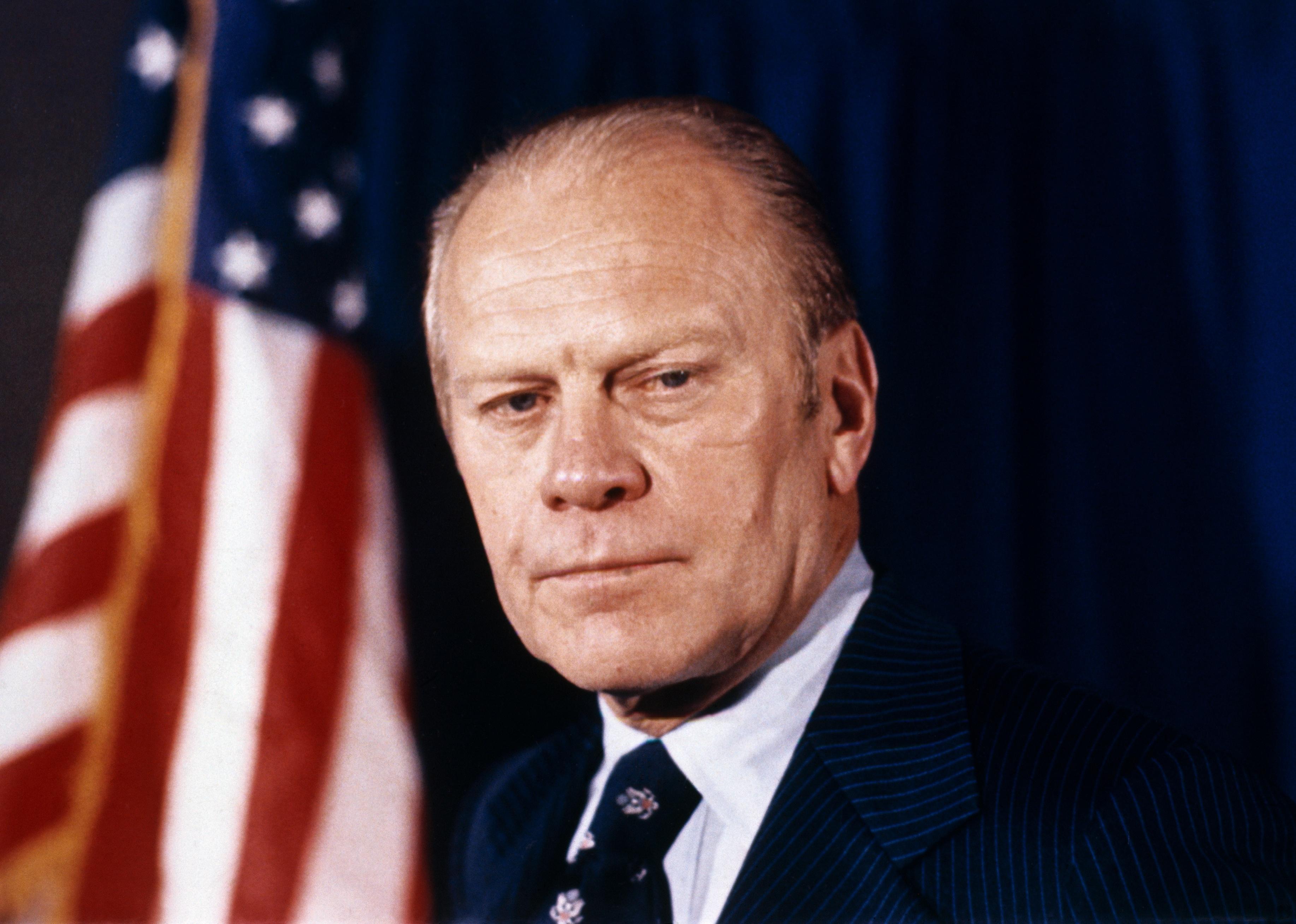 Gerald Ford in a blue pinstripe suit in front of an American flag.