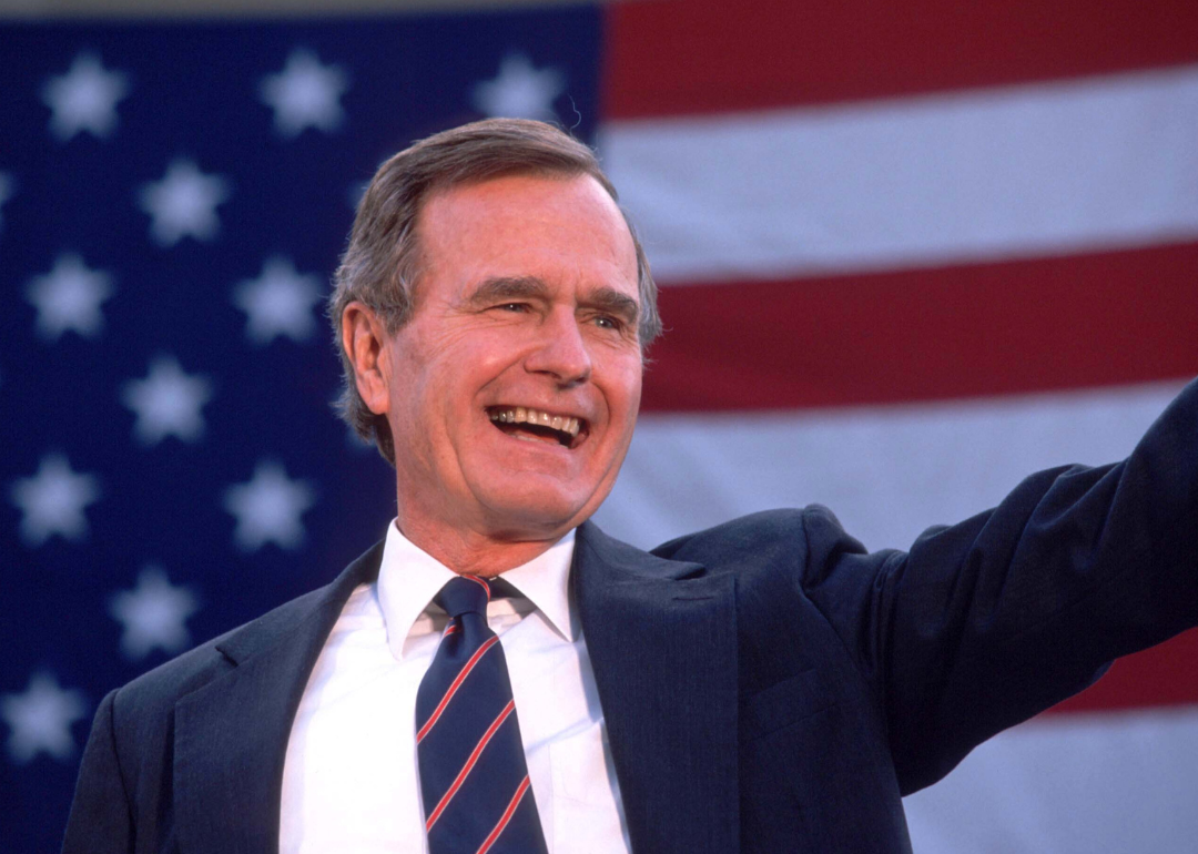 George H.W. Bush waving onstage in a blue suit in front of an American flag.