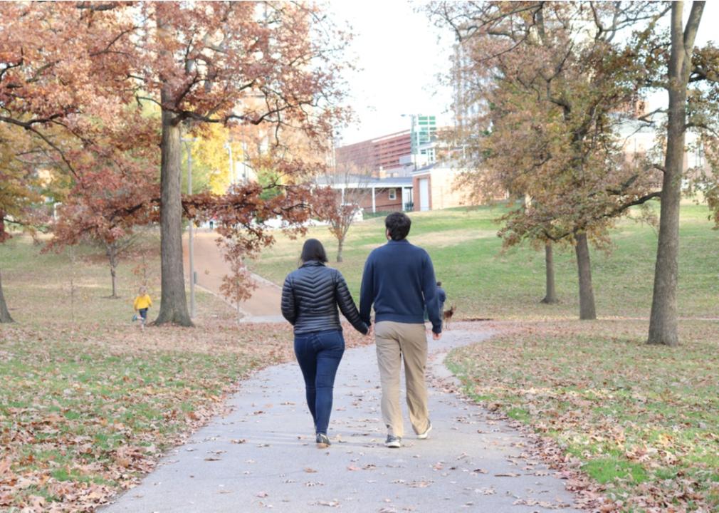 A couple holding hands walking along a path in a park while a child runs in the grass and a man walks his dog ahead.