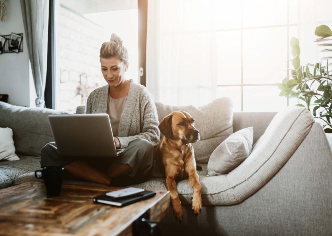 A person sitting on a couch next to a dog while they works on their laptop.