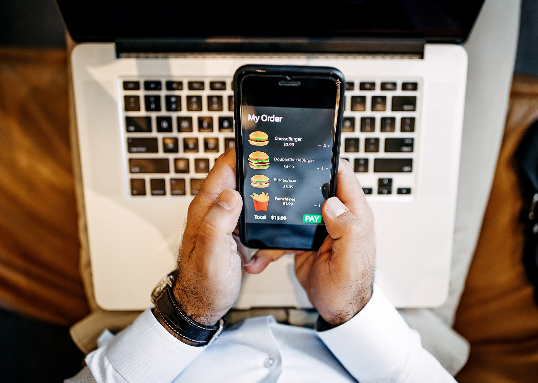 A person in business attire in front of a laptop orders burgers and fries on a cellphone app.