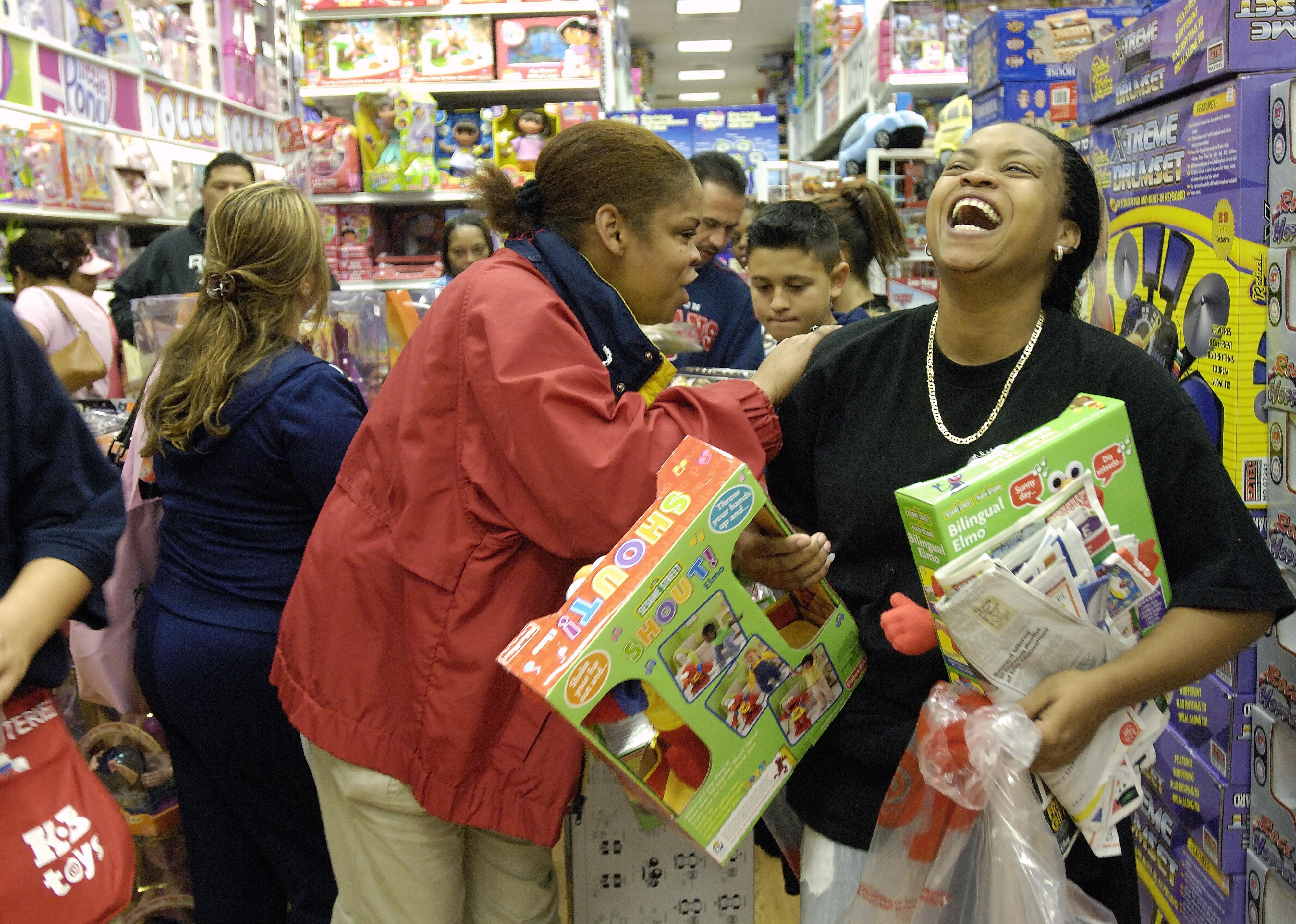 Two women laughing while holding toys in a crowded toy store.