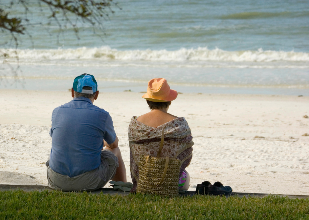 An older man in a ball cap and a woman in a sun hat sit on the edge of some green grass looking down at the beach.