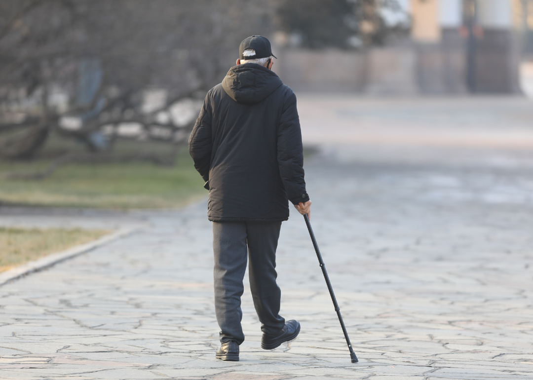 An older man wearing a jacket and walking down a stone road with a cane.