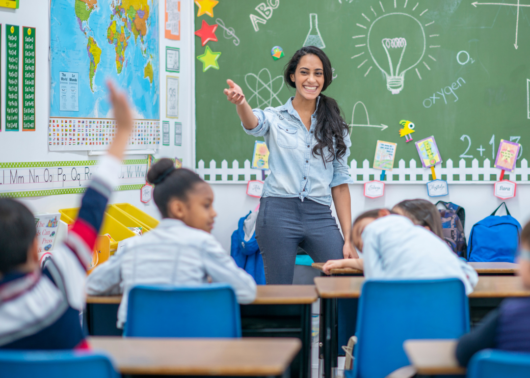 A teacher smiles and motions to a student with his hand raised.