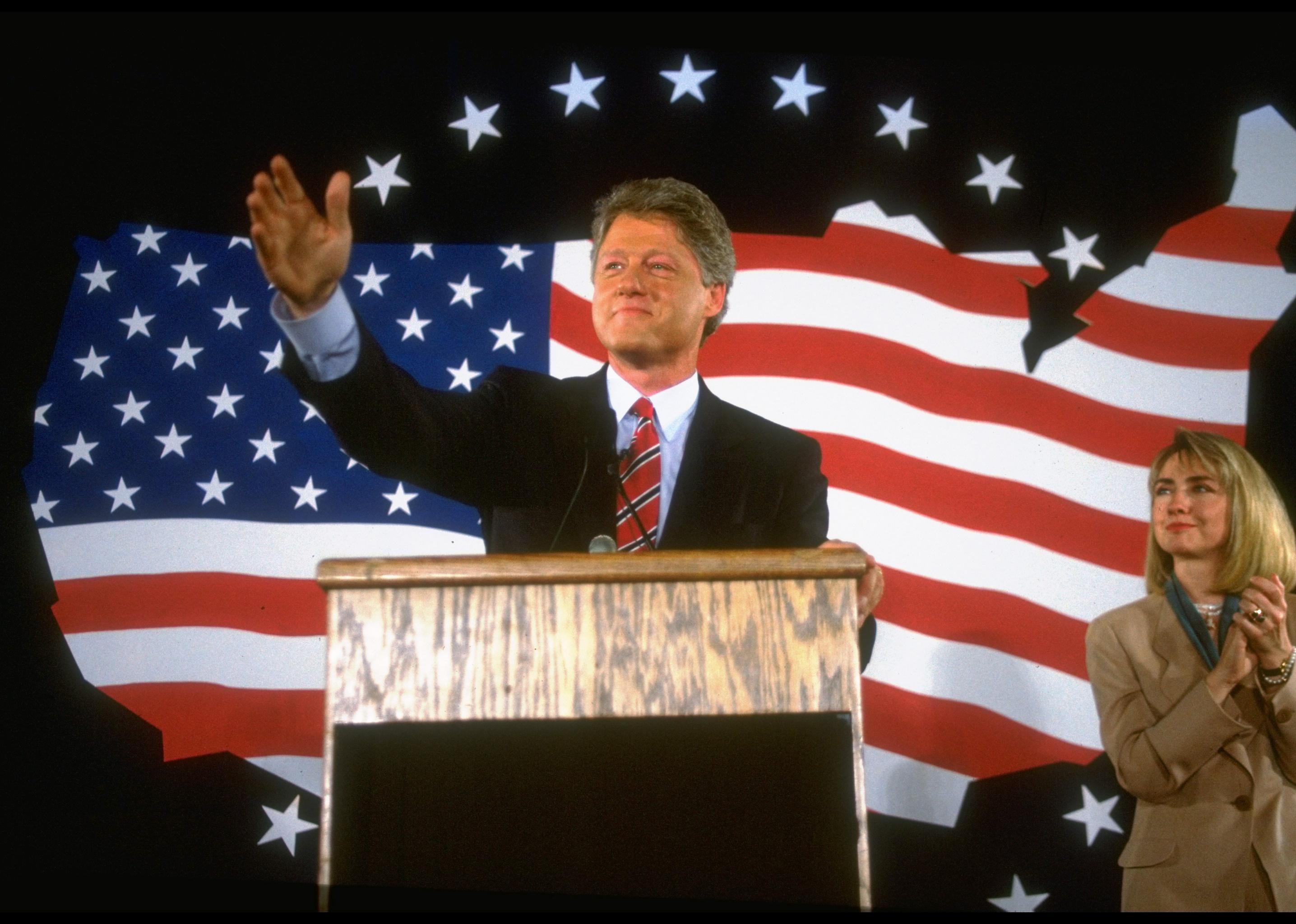 Bill Clinton campaigning for president onstage with Hillary Clinton and an American flag in the background.
